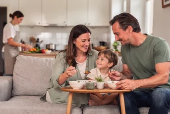 A family eating together on the sofa while a chef cooks in their kitchen